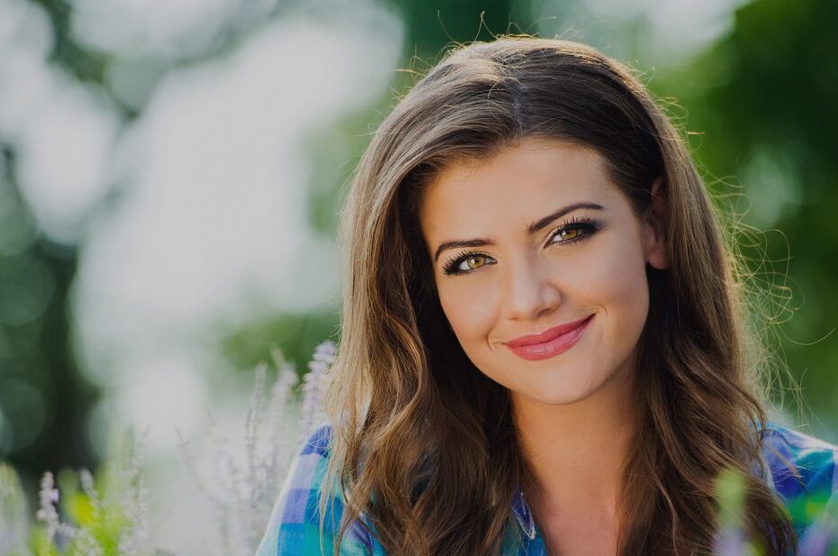 close up of a woman relaxed smiling with blurry green leaves in the background representing wellness.